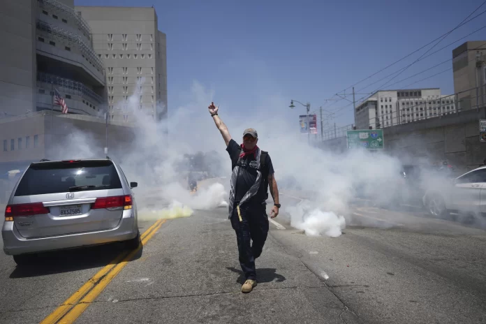 A protester displays a poster as tear gas is used in the metropolitan detention center of downtown Los Angeles, Sunday, June 8, 2025, following last night's immigration raid protest. (AP Photo/Eric Thayer)