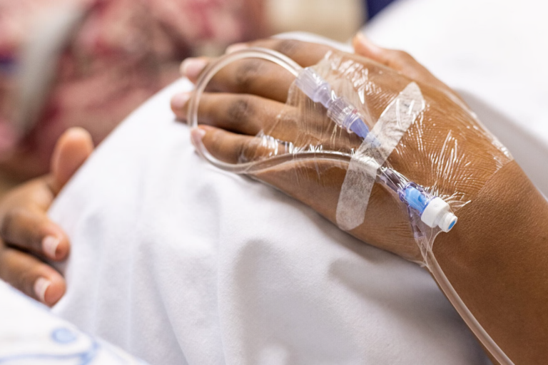 Close-up of a Black woman’s hand resting on a hospital bed with an IV line inserted, highlighting medical care during childbirth or maternal treatment.