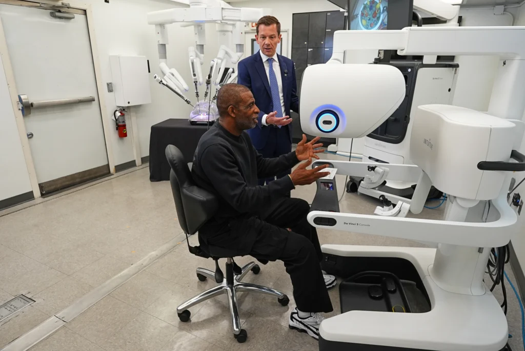 Colorado head football coach Deion Sanders, front, reacts after working the Da Vinci robotic surgical machine used to remove his cancerous bladder last year during a demonstration of the device by Jason Hart, senior vice president of global marketing for Intuitive, Tuesday, June 23, 2026, in Boulder, Colo. (AP Photo/David Zalubowski)